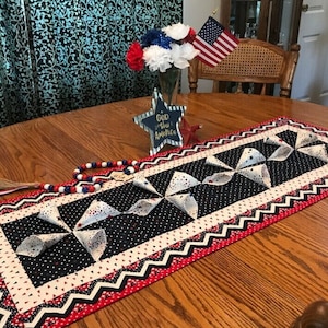 May include: A red, white, and blue table runner with a patriotic theme. The runner features a black background with white polka dots and red hearts. The runner has a white border with red and black zig-zag trim. The runner is decorated with white pinwheels with red and blue accents. The runner is on a wooden table with a wooden chair and a vase of red and white flowers in the background. The text "God Bless America" is on a wooden star.