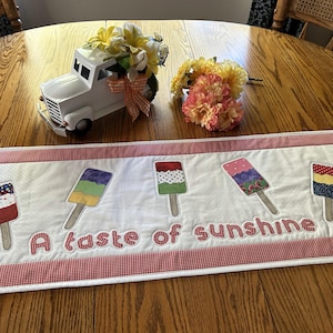 May include: A white table runner with a red gingham border features embroidered popsicle designs in various colors. The text "A taste of sunshine" is embroidered in red. A white truck with flowers sits on the table.