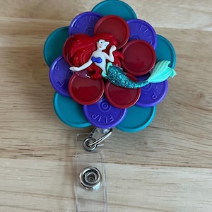 May include: A badge reel featuring a mermaid figurine atop a flower-shaped design. The flower is composed of teal, purple, and red circular elements. The mermaid has red hair, a blue top, and a green tail. A clear plastic badge holder and metal clip are attached.
