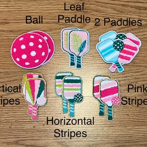 May include: A collection of embroidered patches featuring pickleball equipment. Designs include pink polka dot balls, leaf paddles, and paddles with vertical and horizontal stripes in various colors. Text labels identify each design.