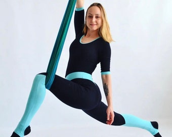 May include: A woman in a black and blue jumpsuit is performing aerial yoga in a blue and green hammock. She is holding the hammock with her right hand and her left leg is extended to the side.