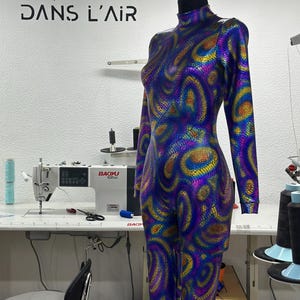May include: A mannequin displaying a full-body jumpsuit with a high neck and long sleeves. The fabric has an iridescent pattern in blue, purple, and yellow. The text "DANS L'AIR" is visible on the wall behind the mannequin.