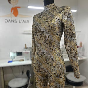 May include: A patterned jumpsuit in gold, silver, and black, displayed on a mannequin. The jumpsuit has a high neck and long sleeves. The background shows a sewing machine and the text "DANS L'AIR".