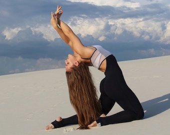 May include: A woman in a white and black one-piece outfit performs a yoga pose on a white sandy beach. The sky is blue with white clouds.