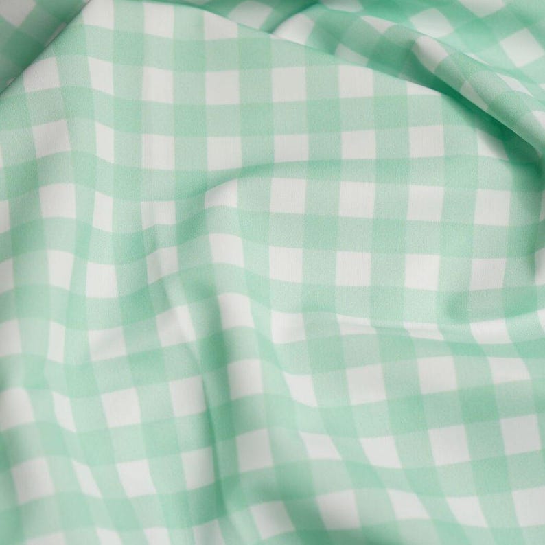 K&ouml;nnte beinhalten: Ein Stoff mit einem klassischen Vichy-Karo-Muster, bestehend aus abwechselnden Quadraten in sanftem Mintgr&uuml;n und klarem Wei&szlig;. Das Material ist in Falten gelegt, wodurch das gleichm&auml;&szlig;ige Karomuster auf der gesamten Textiloberfl&auml;che sichtbar wird.