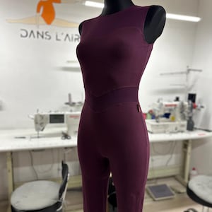 Könnte beinhalten: Ein pflaumenfarbener Jumpsuit auf einer Schaufensterpuppe. Das ärmellose Kleidungsstück hat einen Rundhalsausschnitt, ein figurbetontes Oberteil mit Chevron-Design und eine stromlinienförmige Silhouette. Der Jumpsuit wird in einem Studio mit Nähutensilien und dem Text "DANS L'AIR" im Hintergrund gezeigt.