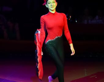 May include: A performer in a red and black bodysuit with sparkling accents and a matching red headpiece. The long-sleeved outfit has a high neckline and is paired with black leggings. The performer is on a red stage, holding a red scarf.
