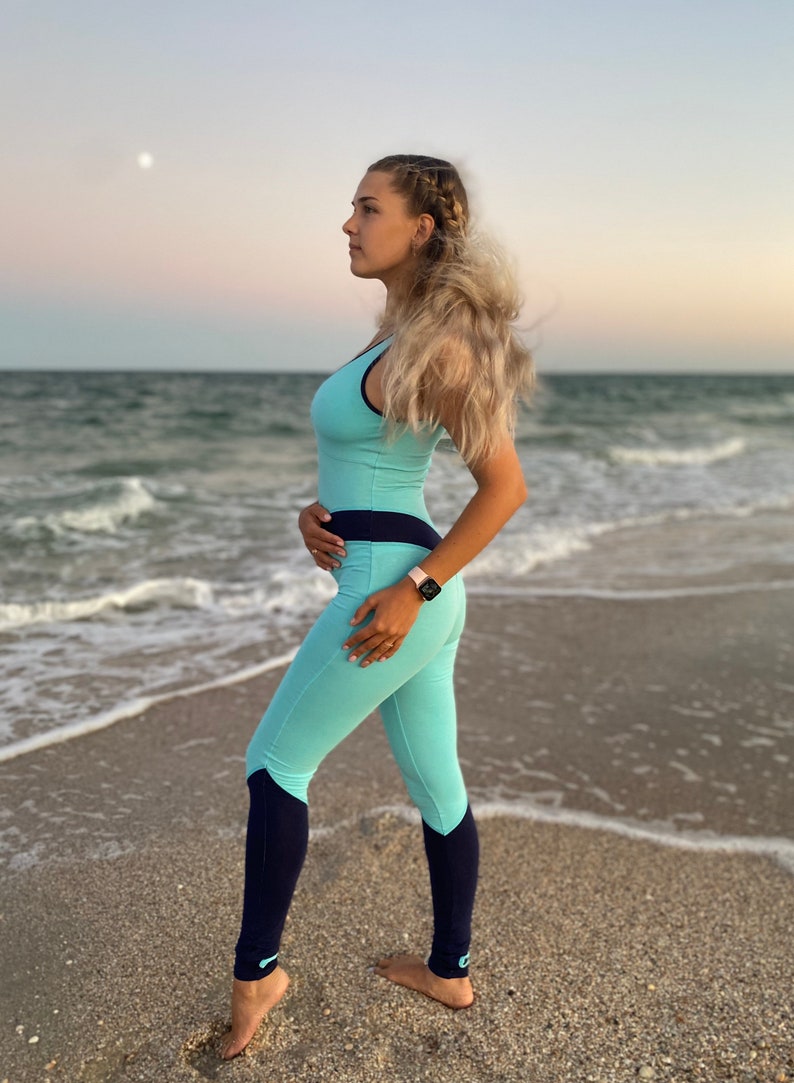 K&ouml;nnte beinhalten: Ein hellblauer und marineblauer Sport-Jumpsuit mit &auml;rmellosem Oberteil, einem breiten marineblauen Taillenbund und einer langen Hose mit marineblauen Beinabschl&uuml;ssen sowie kleinen hellblauen Akzenten an den Kn&ouml;cheln.