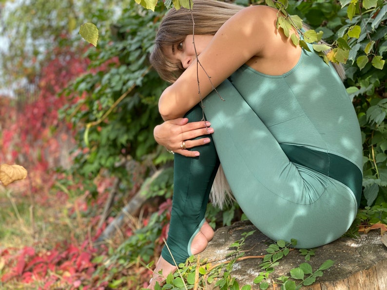 May include: A woman wearing a green one-piece outfit sits on a tree stump in a forest setting. The outfit has a dark green panel on the lower legs and a light green top.