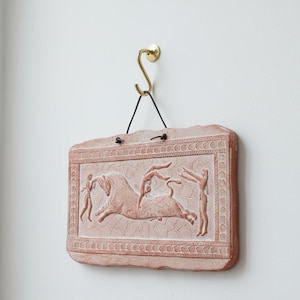 May include: A rectangular, reddish-brown wall plaque with a relief depicting figures and a bull. The plaque is suspended by a black cord from a brass hook. The relief features a border and detailed figures.