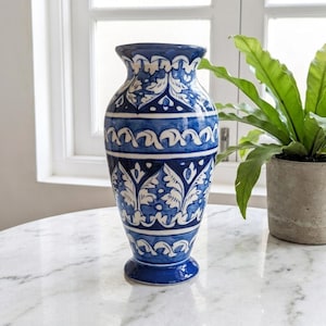 Puede incluir: Un jarrón de cerámica con un patrón pintado a mano en azul y blanco de hojas estilizadas y bandas geométricas. El recipiente tiene un borde acampanado, un cuello estrecho y un cuerpo redondeado que se estrecha hacia una base circular brillante.