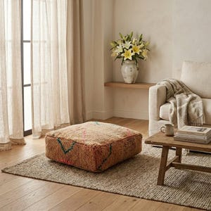 May include: A square, brown floor pouf with colorful geometric patterns. The pouf sits on a neutral-toned rug in a living room setting. A vase of white and yellow flowers sits on a shelf in the background.