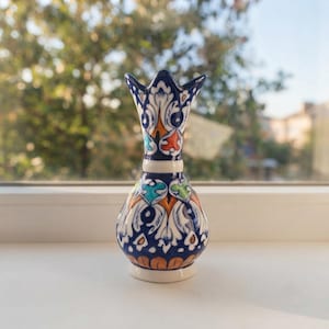 May include: A small ceramic vase with a flared, scalloped rim and a bulbous base. The surface features intricate hand-painted patterns in deep blue, white, orange, and turquoise, showcasing symmetrical floral and leaf-like motifs throughout.