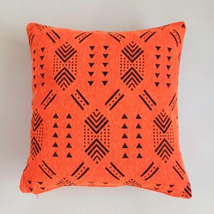 Puede incluir: Cojín decorativo cuadrado naranja con un patrón geométrico negro. El patrón incluye triángulos, líneas y formas punteadas. El cojín tiene una apariencia texturizada y un cierre de cremallera.