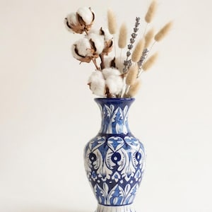 May include: A ceramic vase featuring a deep blue and white intricate floral pattern. The vase is filled with a dried floral arrangement consisting of white cotton bolls, light brown bunny tail grass, and sprigs of purple lavender stems.