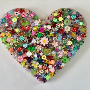 May include: A heart-shaped resin plaque filled with a dense arrangement of colorful beads and charms. The surface features various shapes including flowers, smiley faces, fruits, stars, and animals in a wide spectrum of vibrant colors.