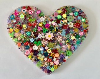 May include: A heart-shaped resin plaque filled with a dense arrangement of colorful beads and charms. The surface features various shapes including flowers, smiley faces, fruits, stars, and animals in a wide spectrum of vibrant colors.
