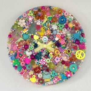 May include: A circular resin coaster embedded with a dense assortment of colorful beads and charms, including a central pale yellow bow, a yellow duck, a blue smiley face, a peace sign, various flowers, stars, hearts, and miniature food items.