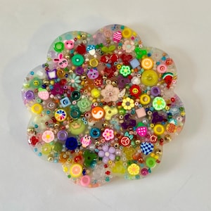 May include: A flower-shaped coaster made of clear resin embedded with a dense collection of colorful beads and charms. The embedded items include miniature fruits, animals, smiley faces, flowers, stars, and geometric shapes in various colors.