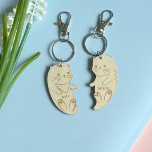 May include: Two wooden otter keychains with silver-colored clasps and key rings. Each otter is engraved with a name, "Amy" and "Brian". The keychains are light brown and feature a heart design. The background is light blue.