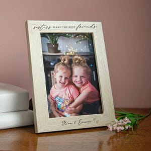 Puede incluir: Un marco de fotos de madera de color claro con el texto "sisters make the best friends" y los nombres "Olivia & Emma" debajo de la foto. El marco contiene una foto de dos niñas pequeñas abrazándose, con camisas rosas.