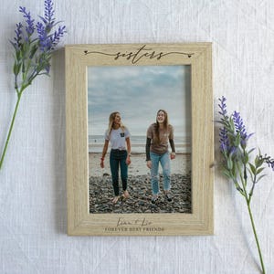 Puede incluir: Un marco de fotos de madera de color claro con la palabra "sisters" en escritura cursiva en la parte superior. El marco contiene una foto de dos mujeres jóvenes caminando por una playa. El texto "Lena & Liz FOREVER BEST FRIENDS" está en la parte inferior.