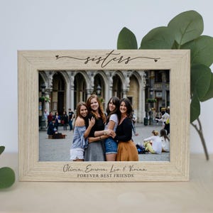 Puede incluir: Un marco de fotos de madera de color claro con la palabra "sisters" en escritura cursiva. El marco contiene una foto de cuatro mujeres. Los nombres Olivia, Emma, Liz & Vivian están debajo de la foto, con "FOREVER BEST FRIENDS" debajo.