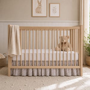May include: A light wood baby cot with a white and beige polka dot fitted sheet and a brown gingham cot skirt. A beige teddy bear sits on the mattress. A beige blanket hangs over the side. Two framed pictures hang on the wall.