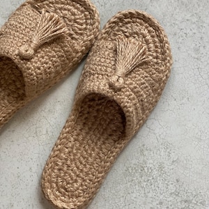 May include: A pair of beige crocheted slippers with tassels. The slippers are made from a natural fibre and have a simple, minimalist design.