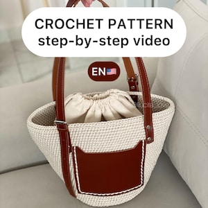 May include: A cream-colored crochet tote bag with brown leather handles and accents. The bag features a brown leather pocket and a drawstring closure. Text on the image reads "CROCHET PATTERN step-by-step video".