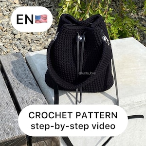 以下が含まれることがあります： 黒いクロシェの巾着バッグに、黒い革の紐とストラップが付いています。バッグは灰色のコンクリートの表面に木製のベンチの上に置かれています。テキスト「CROCHET PATTERN step-by-step video」は、黒い背景に白い文字で書かれています。