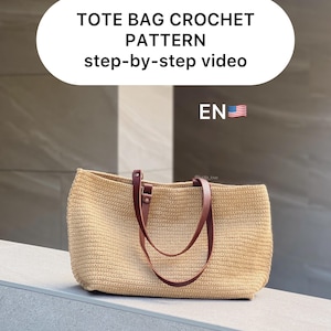 May include: A beige crochet tote bag with brown leather straps. The bag is sitting on a light beige surface. The text "TOTE BAG CROCHET PATTERN step-by-step video EN" is visible in the image.