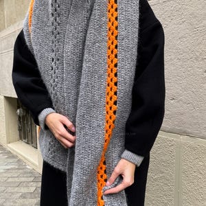 Op de afbeelding: Een grijze sjaal met een oranje gehaakte streep aan de zijkant. De sjaal is gemaakt van een zacht, getextureerd materiaal. De sjaal is om de schouders gewikkeld.
