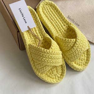 Może przedstawiać: Para żółtych, szydełkowych kapci z krzyżującym się paskiem. Kapcie są oznaczone "Liucita Love".