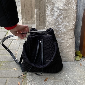 Könnte beinhalten: Ein schwarzer Häkelrucksack mit Kordelzugverschluss und Lederriemen. Die Tasche hat ein strukturiertes Aussehen und ein schlichtes, modernes Design. Der Rucksack wird im Freien gezeigt.