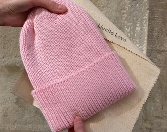 Könnte beinhalten: Eine rosafarbene Strickmütze mit gerippter Struktur. Die Mütze wird von einer Hand gehalten und liegt auf einem beigefarbenen Stoff mit dem aufgedruckten Text "Liucita Love".