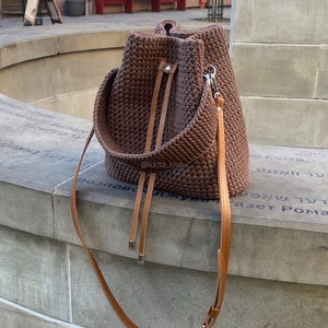 May include: A brown, handmade crochet bucket bag with a leather shoulder strap and drawstring closure. The bag has a wide, braided top handle and a silver-tone clasp. The bag is sitting on a stone ledge.