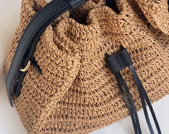 May include: A brown woven straw bucket bag with a black leather strap and drawstring closure. The bag has a gold metal accent on the strap.