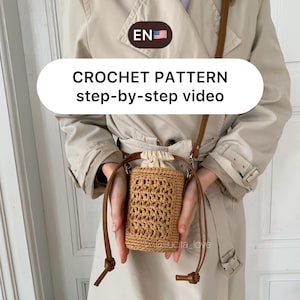 May include: A crocheted bag with a brown leather strap. The bag is being held by a person wearing a beige trench coat. The text "CROCHET PATTERN step-by-step video" is displayed on a white circle.
