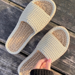 Op de afbeelding: Een paar crèmekleurige slippers met open teen. De slippers hebben een geweven textuur aan de bovenkant en een voetbed van natuurlijke vezels. De randen zijn afgewerkt met een lichter crèmekleurig materiaal. De slippers liggen op een houten oppervlak.