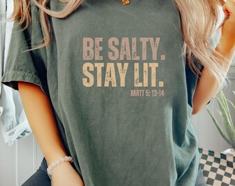 May include: A dark green t-shirt with the text "BE SALTY. STAY LIT." in a distressed font. The text "MATT 5:13-14" is printed in a smaller font below the main text.