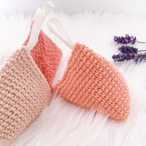 May include: Two crocheted triangular sachets in shades of pink and peach, with white ribbon ties. The sachets are sitting on a white fluffy surface.