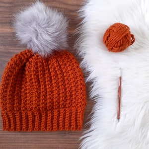 May include: A crocheted beanie hat in a burnt orange color with a gray faux fur pom pom. The hat is lying on a wooden surface. A ball of orange yarn, a crochet hook, and a white faux fur rug are in the background.