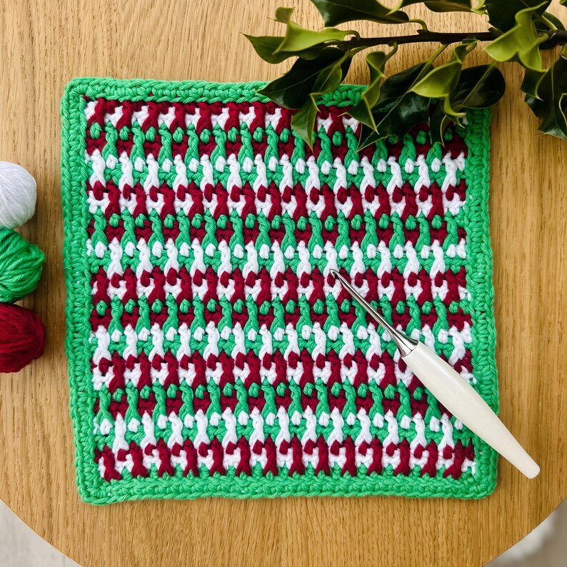 May include: A green, red, and white crocheted square with a green border. A crochet hook is lying on top of the square.