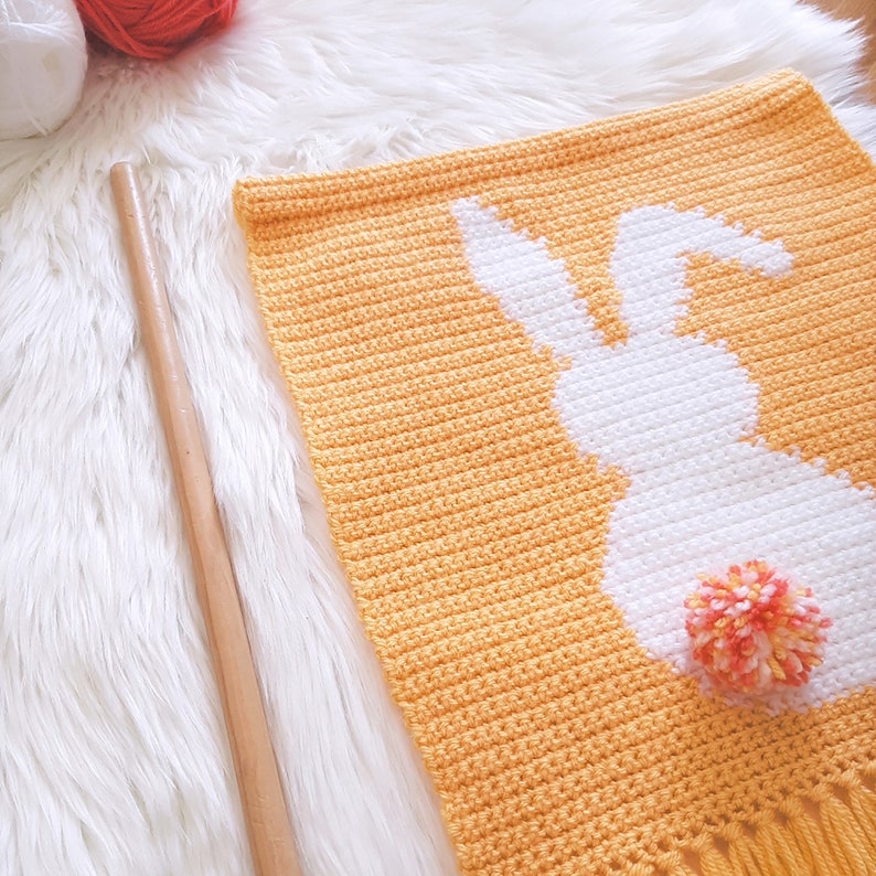 May include: A crocheted wall hanging featuring a white bunny silhouette with a pink and orange pom-pom tail on a yellow background.