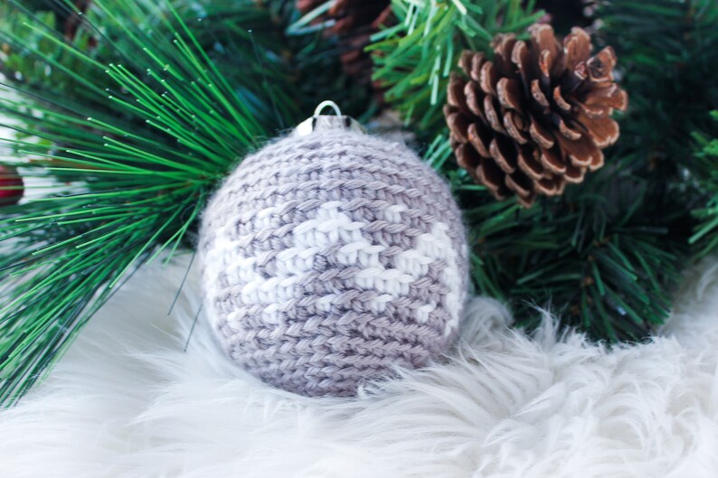 May include: A spherical crochet ornament featuring a light brown and white geometric pattern. The round, textured yarn decoration has a silver-colored metal cap at the top for hanging, showcasing intricate knit stitches throughout the surface.