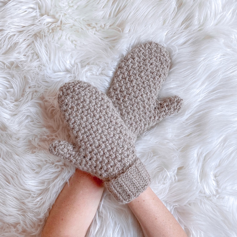 May include: A pair of brown crocheted mittens with a textured stitch pattern. The mittens are laid out on a white fluffy surface.