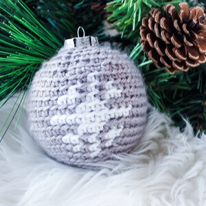 May include: A spherical Christmas ornament featuring a light brown crocheted exterior with a white snowflake pattern stitched into the center. The ornament is topped with a silver-colored metal cap and a small wire loop for hanging.