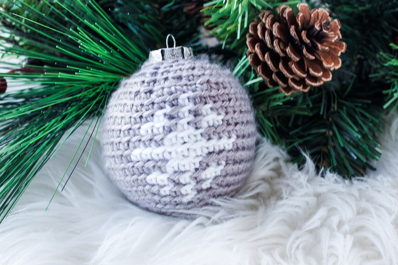 May include: A spherical Christmas ornament featuring a light brown crocheted exterior with a white snowflake pattern stitched into the center. The ornament is topped with a silver-colored metal cap and a small wire loop for hanging.