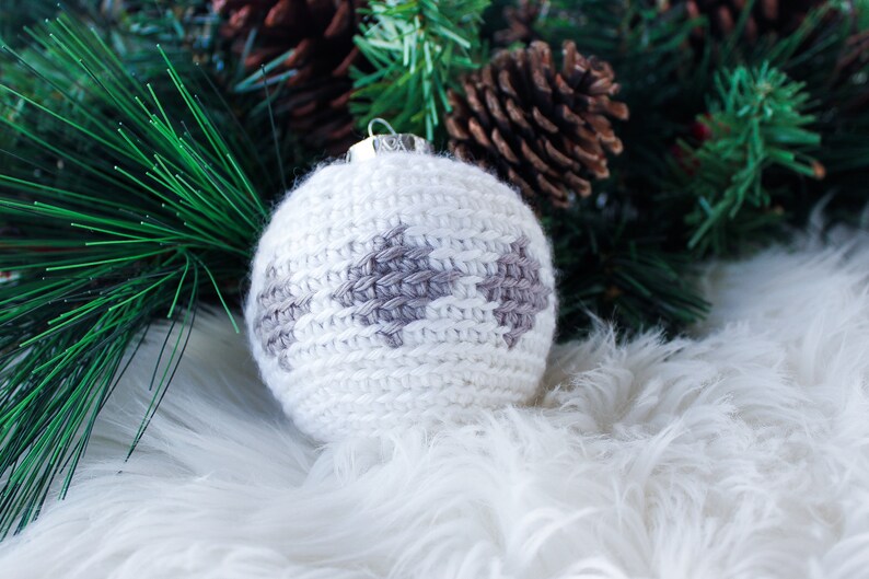 May include: A spherical Christmas ornament featuring white yarn crochet work with a repeating gray diamond pattern. The ornament has a silver-colored metal cap at the top for hanging, showcasing a textured, handmade knit construction.
